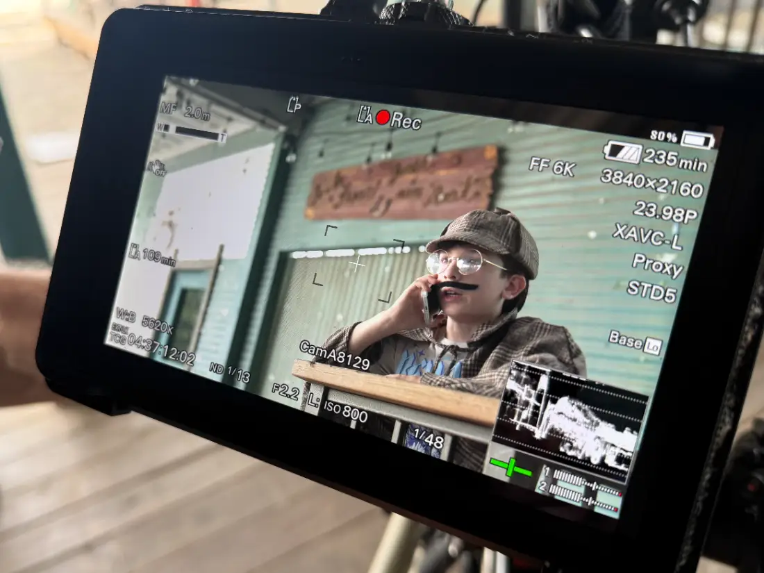 A young actor in a flat cap and moustache on a phone in character, framed up on the camera's monitor mid-take